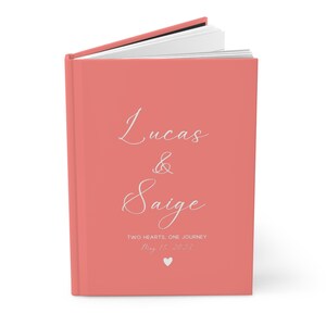 K&ouml;nnte beinhalten: Ein korallenfarbenes Hardcover-G&auml;stebuch mit dem Text "Lucas & Saige" in wei&szlig;er Schrift. Der Text "Two Hearts, One Journey" und das Datum "May 15, 2023" sind ebenfalls in wei&szlig;er Schrift.