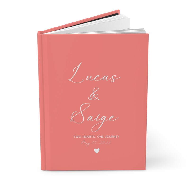 K&ouml;nnte beinhalten: Ein korallenfarbenes Hardcover-G&auml;stebuch mit dem Text "Lucas & Saige" in wei&szlig;er Schrift. Der Text "Two Hearts, One Journey" und das Datum "May 15, 2023" sind ebenfalls in wei&szlig;er Schrift.