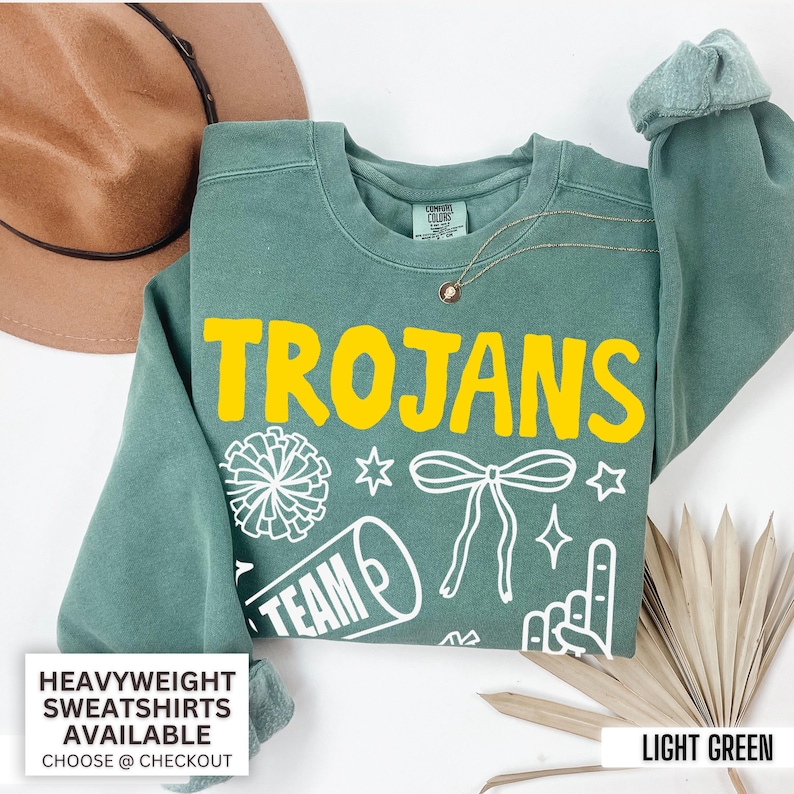 Puede incluir: Una sudadera de cuello redondo en color verde salvia con la palabra TROJANS en letras amarillas llamativas. Debajo del texto hay gr&aacute;ficos de l&iacute;neas blancas de un pomp&oacute;n, un meg&aacute;fono con la palabra TEAM, un lazo, estrellas y un dedo de espuma.