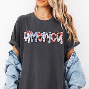May include: A dark gray t-shirt with the word "America" written in white, red, and blue patterned letters. The letters are decorated with red and blue bows.