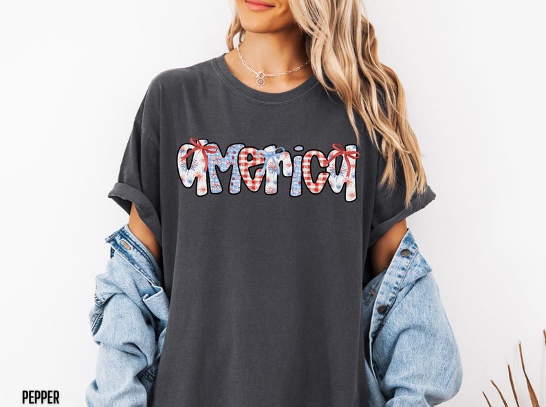 May include: A dark gray t-shirt with the word "America" written in white, red, and blue patterned letters. The letters are decorated with red and blue bows.