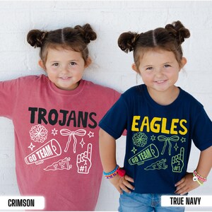 Puede incluir: Una camiseta carmes&iacute; con texto negro que dice TROJANS y gr&aacute;ficos de porristas en blanco, y una camiseta azul marino con texto amarillo que dice EAGLES y gr&aacute;ficos en verde, incluyendo pompones, meg&aacute;fonos, lazos y dedos de espuma.
