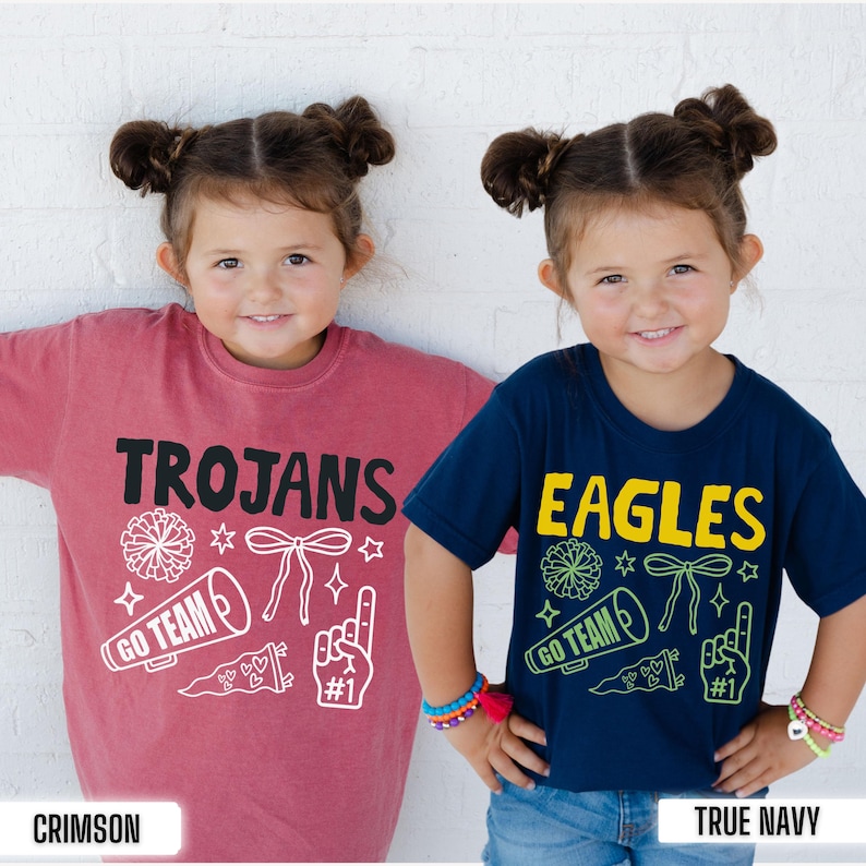 Puede incluir: Una camiseta carmes&iacute; con texto negro que dice TROJANS y gr&aacute;ficos de porristas en blanco, y una camiseta azul marino con texto amarillo que dice EAGLES y gr&aacute;ficos en verde, incluyendo pompones, meg&aacute;fonos, lazos y dedos de espuma.