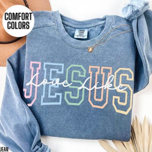 May include: A blue jean sweatshirt featuring the word "Jesus" in rainbow colours with "love like" written in white cursive underneath. The sweatshirt is made from a soft, comfortable material and has a distressed look.