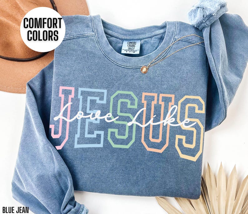 May include: A blue jean sweatshirt featuring the word "Jesus" in rainbow colours with "love like" written in white cursive underneath. The sweatshirt is made from a soft, comfortable material and has a distressed look.