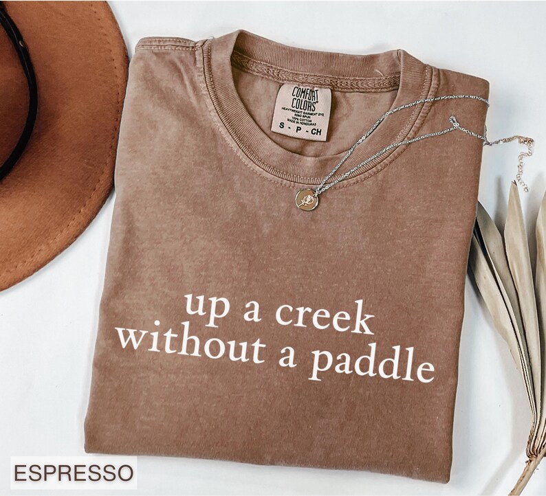 This funny shirt says up a creek without a paddle, just like the southern saying that is said in the south. This would be a funny gag gift for a friend, mom, sister or coworker.  This Comfort Colors shirt comes in 6 beautiful colors, espresso, light green, moss, blue jean, violet and pepper.  This humor tee will make a humorous gift. Share your sarcasm through your sarcastic apparel.
