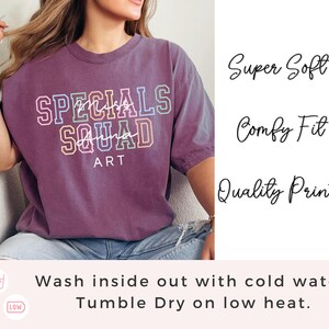 May include: A purple t-shirt featuring the text "SPECIALS SQUAD ART" in a multi-coloured, layered design. The shirt also has the phrases "Super Soft", "Comfy Fit", and "Quality Print" printed on it. The shirt is being worn by a person.