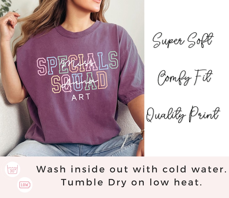May include: A purple t-shirt featuring the text "SPECIALS SQUAD ART" in a multi-coloured, layered design. The shirt also has the phrases "Super Soft", "Comfy Fit", and "Quality Print" printed on it. The shirt is being worn by a person.