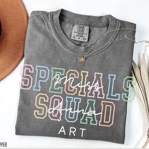 May include: A grey Comfort Colors t-shirt with the words "Specials Squad Art" in colourful block letters. A silver necklace with a pendant rests on the shirt. The shirt is folded and displayed on a white surface.