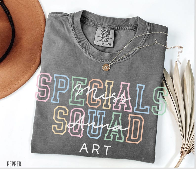May include: A grey Comfort Colors t-shirt with the words "Specials Squad Art" in colourful block letters. A silver necklace with a pendant rests on the shirt. The shirt is folded and displayed on a white surface.