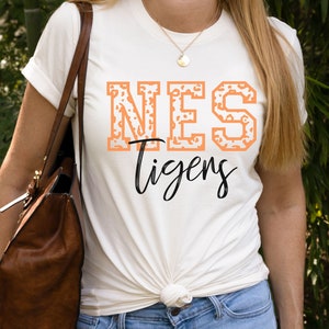 May include: A white t-shirt with a peach-colored "NES Tigers" graphic. The "NES" is in a blocky font with a leopard print pattern.