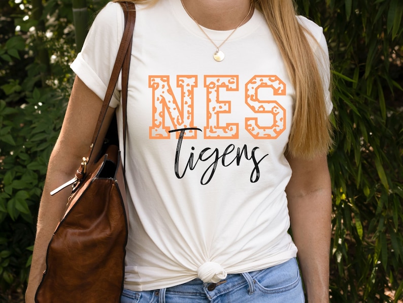 May include: A white t-shirt with a peach-colored "NES Tigers" graphic. The "NES" is in a blocky font with a leopard print pattern.