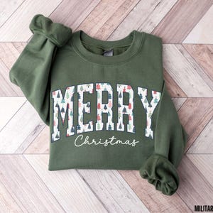 May include: A green sweatshirt with a white "Merry Christmas" graphic featuring a pattern of Christmas trees. The text is in a collegiate font.
