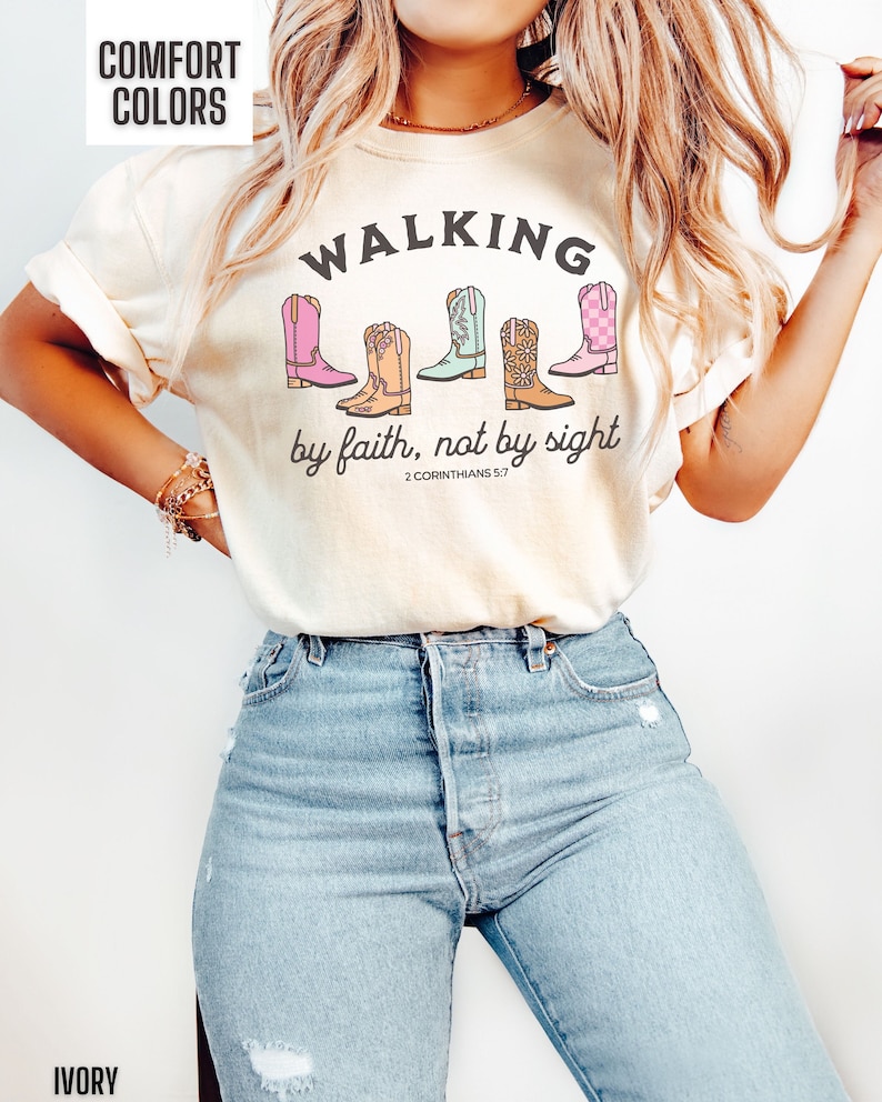 Puede incluir: Camiseta de color marfil con el texto "WALKING by faith, not by sight" y una referencia b&iacute;blica. La camiseta presenta un gr&aacute;fico de cinco botas de vaquero de colores.