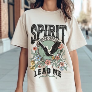 K&ouml;nnte beinhalten: Ein cremefarbenes T-Shirt mit einem Vintage-Grafikdesign, das einen schwarzen Vogel mit ausgebreiteten Fl&uuml;geln, einen gr&uuml;nen Kreis und florale Akzente zeigt. Der Text "SPIRIT LEAD ME" ist in Schwarz gedruckt.