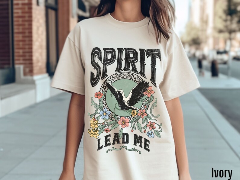 K&ouml;nnte beinhalten: Ein cremefarbenes T-Shirt mit einem Vintage-Grafikdesign, das einen schwarzen Vogel mit ausgebreiteten Fl&uuml;geln, einen gr&uuml;nen Kreis und florale Akzente zeigt. Der Text "SPIRIT LEAD ME" ist in Schwarz gedruckt.