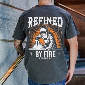 May include: Dark gray t-shirt with a graphic of a welder in a welding mask, with the words "REFINED BY FIRE" and "1 PETER 1:7" in white and orange. The design features a sunburst effect behind the welder.