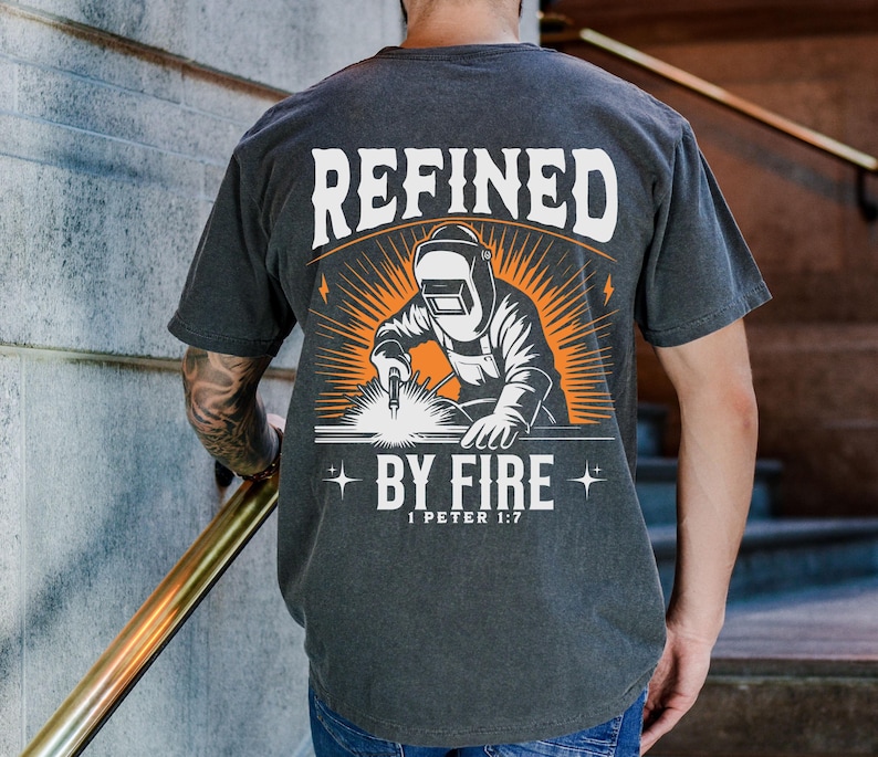 May include: Dark gray t-shirt with a graphic of a welder in a welding mask, with the words "REFINED BY FIRE" and "1 PETER 1:7" in white and orange. The design features a sunburst effect behind the welder.