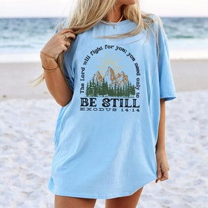 May include: Light blue t-shirt with a graphic of a mountain range and a sun. The text "The Lord will fight for you; you need only to BE STILL EXODUS 14:14" is printed on the shirt.