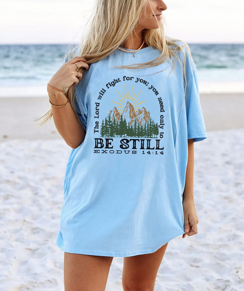 May include: Light blue t-shirt with a graphic of a mountain range and a sun. The text "The Lord will fight for you; you need only to BE STILL EXODUS 14:14" is printed on the shirt.