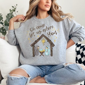 Puede incluir: Una sudadera gris con el texto "Oh come let us adore Him" y una representaci&oacute;n de la escena de la Natividad. El texto est&aacute; en color dorado y la escena de la Natividad est&aacute; en marr&oacute;n y azul.