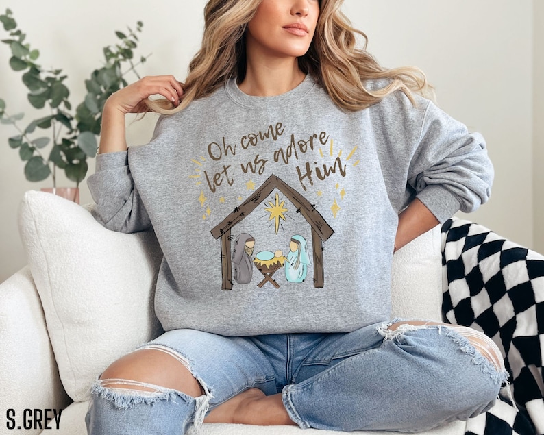 Puede incluir: Una sudadera gris con el texto "Oh come let us adore Him" y una representaci&oacute;n de la escena de la Natividad. El texto est&aacute; en color dorado y la escena de la Natividad est&aacute; en marr&oacute;n y azul.