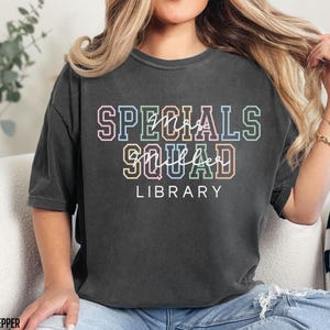 May include: A dark grey t-shirt featuring the text "SPECIALS SQUAD LIBRARY" in a multi-coloured, outlined font. The words are arranged vertically, with "SPECIALS" at the top, "SQUAD" in the middle, and "LIBRARY" at the bottom. The shirt has a relaxed fit.