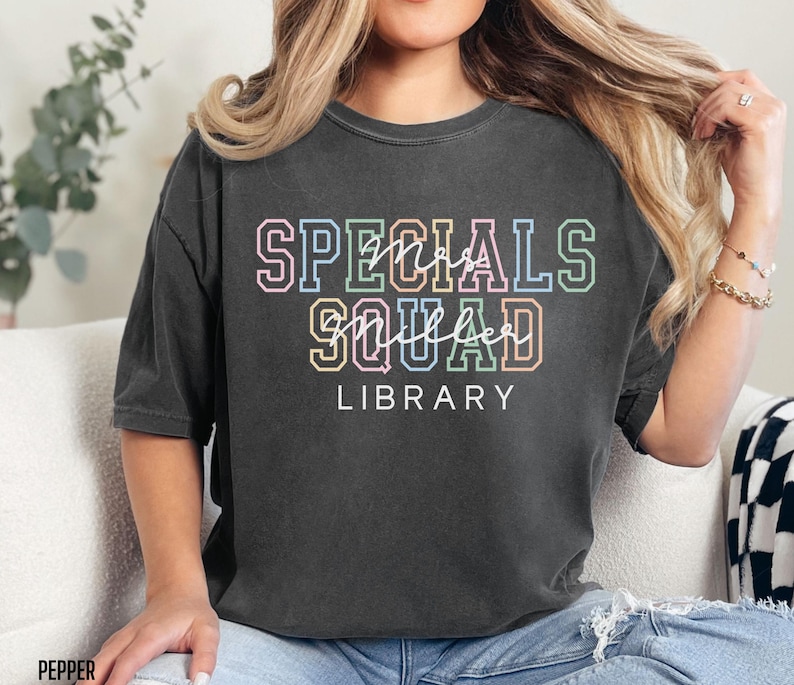 May include: A dark grey t-shirt featuring the text "SPECIALS SQUAD LIBRARY" in a multi-coloured, outlined font. The words are arranged vertically, with "SPECIALS" at the top, "SQUAD" in the middle, and "LIBRARY" at the bottom. The shirt has a relaxed fit.