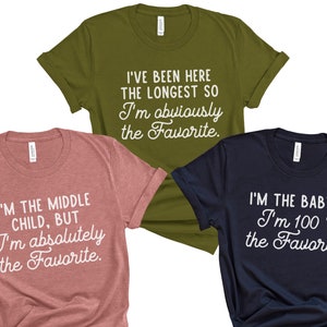 May include: Three t-shirts with different slogans about being the favorite child. The first shirt is olive green and says "I've been here the longest so I'm obviously the favorite." The second shirt is pink and says "I'm the middle child, but I'm absolutely the favorite." The third shirt is navy blue and says "I'm the baby & I'm 100% the favorite."