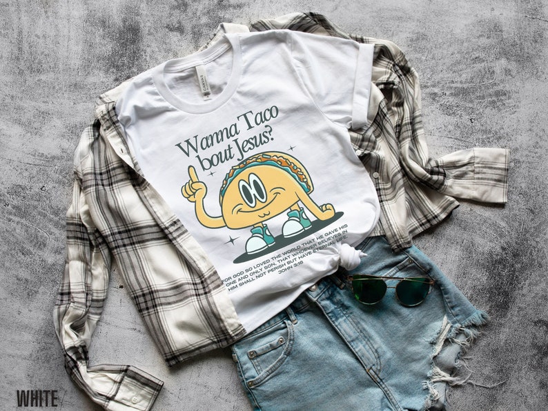 May include: White t-shirt with a cartoon taco graphic and the text "Wanna Taco bout Jesus?"  The taco is wearing sneakers and has a surprised expression. The text "For God so loved the world that He gave His one and only Son, that whoever believes in Him shall not perish but have eternal life. John 3:16" is printed below the graphic.