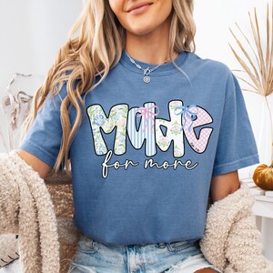 Op de afbeelding: Blauw spijkerbroekkleurig T-shirt met de woorden "MADE for more" in een decoratief lettertype. Het woord "MADE" heeft bloemen-, gestreepte en ruitpatronen. Het shirt is effen van kleur en heeft een relaxte pasvorm.