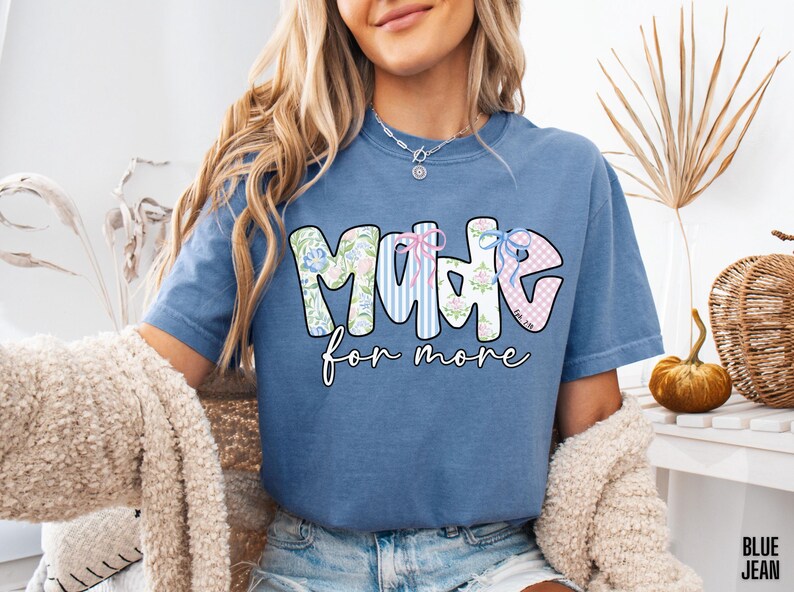 Op de afbeelding: Blauw spijkerbroekkleurig T-shirt met de woorden "MADE for more" in een decoratief lettertype. Het woord "MADE" heeft bloemen-, gestreepte en ruitpatronen. Het shirt is effen van kleur en heeft een relaxte pasvorm.