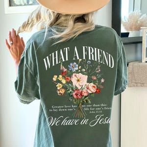 K&ouml;nnte beinhalten: Moosgr&uuml;nes T-Shirt mit dem Text "WHAT A FRIEND" und einem Blumengesteck-Design. Das Shirt zeigt auch den Text "Greater love has no one than this: life for one's friend. We have in Jesus."
