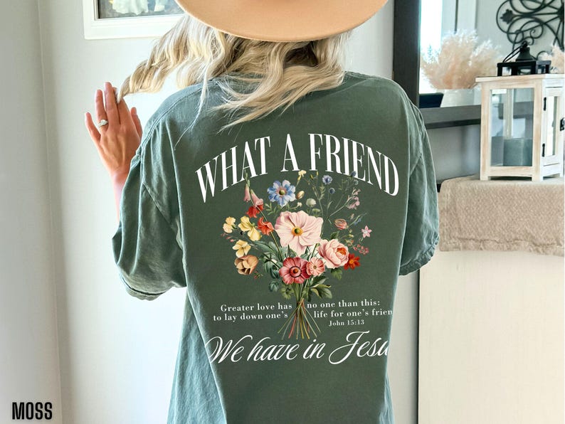 K&ouml;nnte beinhalten: Moosgr&uuml;nes T-Shirt mit dem Text "WHAT A FRIEND" und einem Blumengesteck-Design. Das Shirt zeigt auch den Text "Greater love has no one than this: life for one's friend. We have in Jesus."