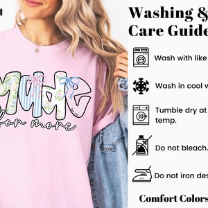 Op de afbeelding: Zachtroze T-shirt met het woord "MADE" in een bubbelletterontwerp, met bloemen-, gestreepte en geruite patronen. De zin "for more" staat eronder. Was- en verzorgingsinstructies staan rechts.