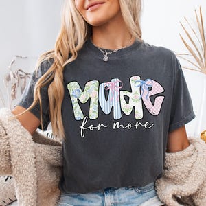 Op de afbeelding: Donkergrijs t-shirt met de woorden "MADE for more" in een kleurrijk, bloemen- en gestreept ontwerp. Het woord "MADE" heeft een bloemenpatroon, strepen en een ruitpatroon. De tekst is in een speels lettertype.
