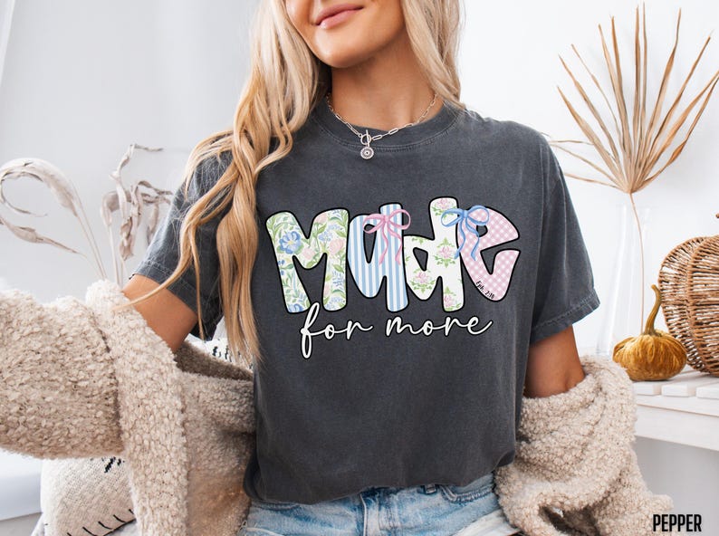 Op de afbeelding: Donkergrijs t-shirt met de woorden "MADE for more" in een kleurrijk, bloemen- en gestreept ontwerp. Het woord "MADE" heeft een bloemenpatroon, strepen en een ruitpatroon. De tekst is in een speels lettertype.