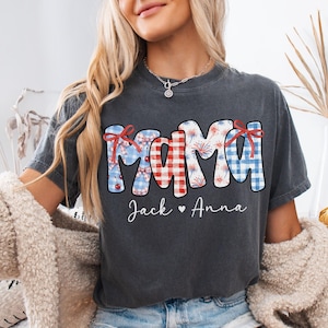 May include: Dark gray t-shirt with the word "MAMA" in red, white, and blue patterns, with the names "Jack" and "Anna" below. The design includes bows and fireworks, perfect for a patriotic theme.