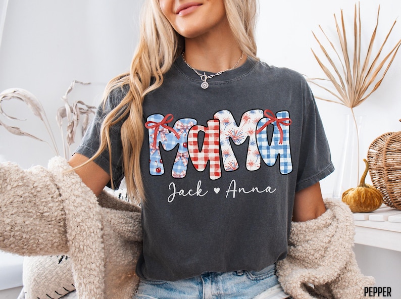 May include: Dark gray t-shirt with the word "MAMA" in red, white, and blue patterns, with the names "Jack" and "Anna" below. The design includes bows and fireworks, perfect for a patriotic theme.
