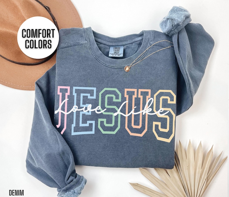 May include: A dark blue sweatshirt featuring the word "JESUS" in rainbow colours and the words "love like" written in white cursive underneath. The sweatshirt is made from a soft, comfortable fabric and has a relaxed fit.