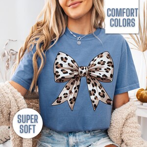 Puede incluir: Una camiseta azul con un dise&ntilde;o de lazo de estampado de leopardo. La camiseta tiene el texto "COMFORT COLORS" y "SUPER SOFT" impreso en ella.