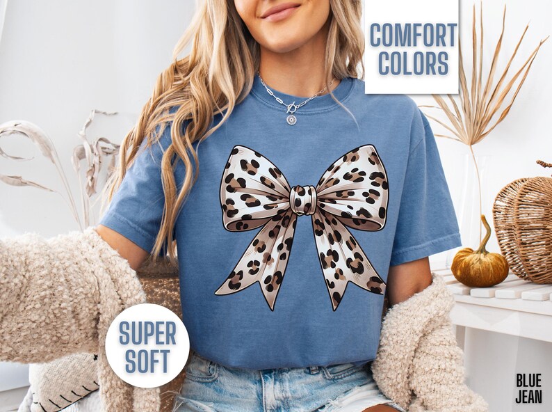 Puede incluir: Una camiseta azul con un dise&ntilde;o de lazo de estampado de leopardo. La camiseta tiene el texto "COMFORT COLORS" y "SUPER SOFT" impreso en ella.