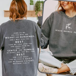 May include: A dark gray sweatshirt with a white graphic print that reads "Come to me. All you who are weary and burdened, and I will give you rest. Take my yoke upon you and learn from me. For I am gentle and humble in heart, and you will find rest for your souls. For my yoke is easy and my burden is light. Matthew 11:28-30". The sweatshirt also has a small white bird graphic on the right side with the text "Jesus brings peace".