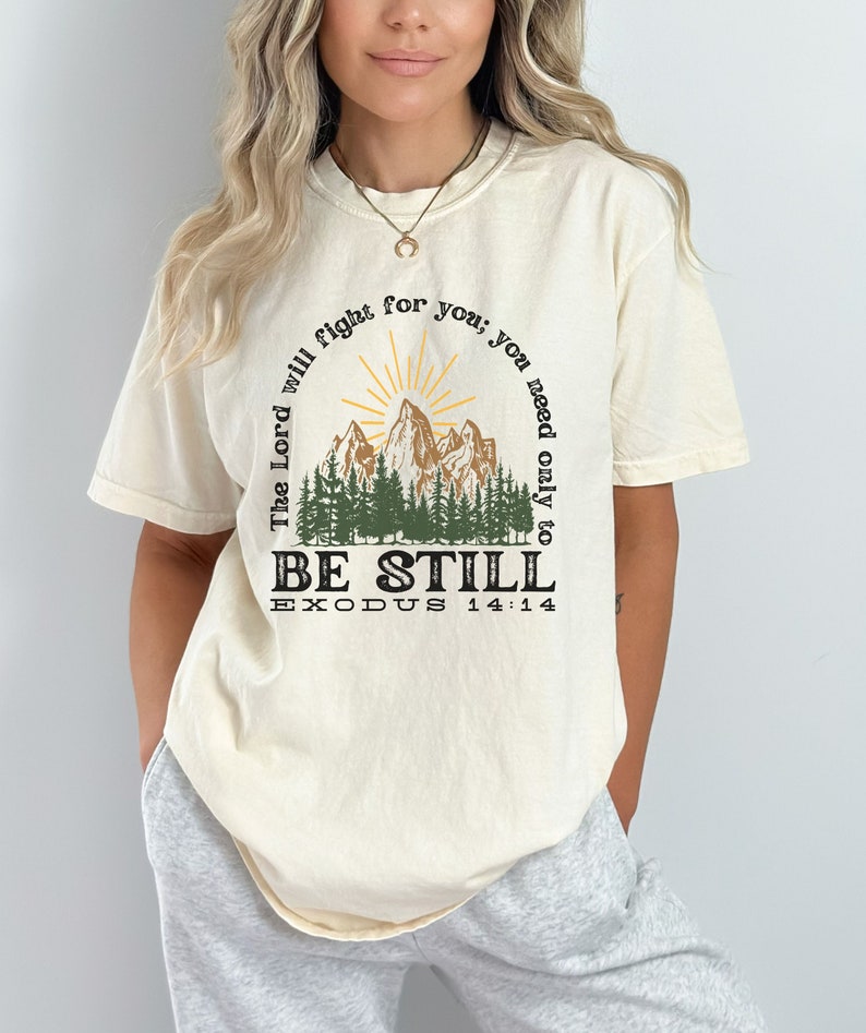 May include: A beige t-shirt with a graphic print of a mountain range with a sun rising behind it. The text "The Lord will fight for you, you need only to be still" is printed in a circle around the mountain range. The text "Exodus 14:14" is printed below the mountain range.