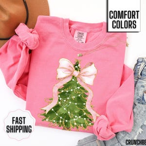 K&ouml;nnte beinhalten: Ein pinkfarbenes Langarm-Sweatshirt mit einem Weihnachtsbaum-Design, einer gro&szlig;en rosa Schleife und goldenen Akzenten. Die Worte "Comfort Colors" und "Fast Shipping" sind sichtbar. Das Sweatshirt wird mit einer blauen Jeans kombiniert.