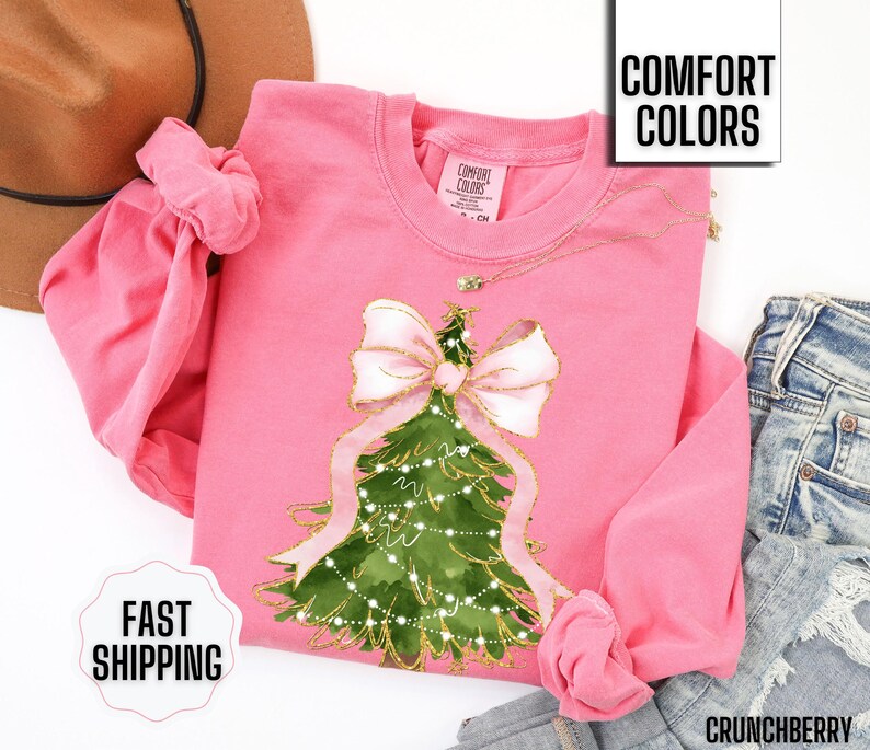 K&ouml;nnte beinhalten: Ein pinkfarbenes Langarm-Sweatshirt mit einem Weihnachtsbaum-Design, einer gro&szlig;en rosa Schleife und goldenen Akzenten. Die Worte "Comfort Colors" und "Fast Shipping" sind sichtbar. Das Sweatshirt wird mit einer blauen Jeans kombiniert.