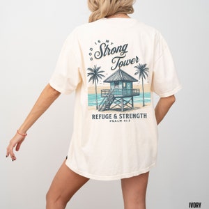 Puede incluir: Una camiseta blanca con un gr&aacute;fico de una casa de playa con palmeras y el texto "God is my Strong Tower Refuge & Strength Psalm 61:3".