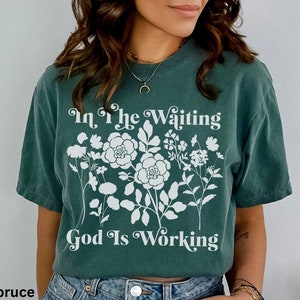 Op de afbeelding: Een tealblauw t-shirt met een wit bloemenmotief en de tekst "In the Waiting God Is Working" gedrukt op de voorkant.
