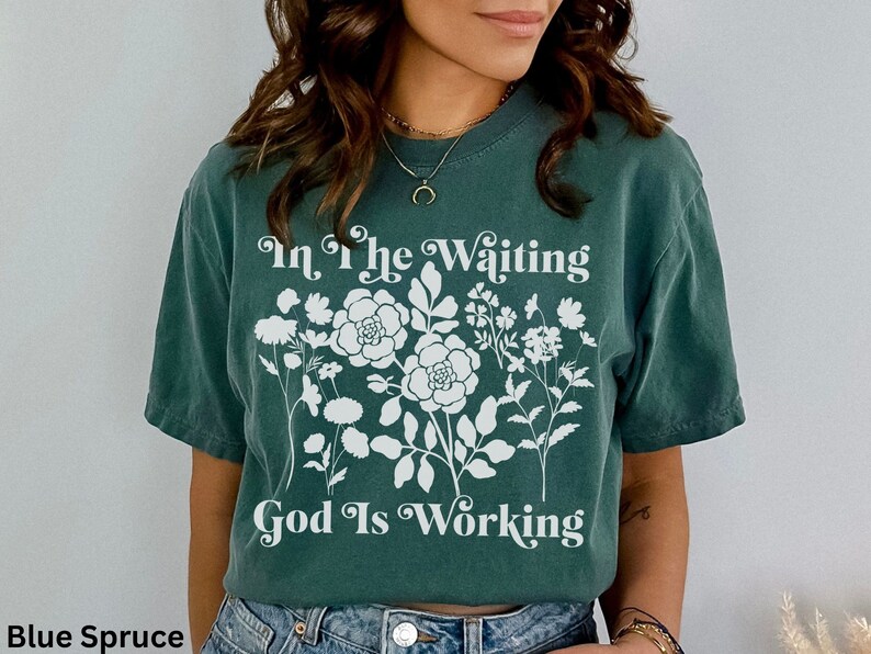 Op de afbeelding: Een tealblauw t-shirt met een wit bloemenmotief en de tekst "In the Waiting God Is Working" gedrukt op de voorkant.
