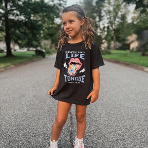 Pu&ograve; includere: Una maglietta nera con la scritta "DEATH AND LIFE" sopra una grafica di una bocca e una lingua piena di fiori. Il testo "TONGUE" e "Prov. 18:21" sono sotto. La maglietta &egrave; indossata da una bambina.